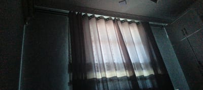 The image depicts a close-up, low-angle view of a window with curtains in what appears to be a room. The curtains are of a sheer, light-colored fabric with a dark, patterned band along the bottom. They are hung from a dark rod and have metal grommets. Light is filtering through the curtains, creating subtle highlights and shadows. The walls on either side of the window are dark and textured, possibly granite or a similar material, with visible speckling. A faint, almost imperceptible square object is visible on the ceiling in the upper portion of the image, possibly a light fixture or vent. The overall impression is of an indoor scene, possibly a residential or commercial space, with the focus on the window treatment. There are no people or active subjects visible in the image. The specific location in the city of Türkmenbaşy, Turkmenistan cannot be determined from the visual information alone.
