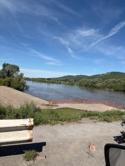 This image depicts a wide river with trees and hills on the opposite bank. The sky is clear with scattered clouds. In the foreground, there is a concrete embankment and a gravel path. A red metal railing runs along the edge of a concrete area near the river. A single person can be seen standing by the railing. In the lower left of the image, a portion of a white barrier with rust marks is visible. A vehicle's side mirror and part of its bumper are in the lower right corner. The location is identified as Nazarovo, Russia, likely along the Chulym River.