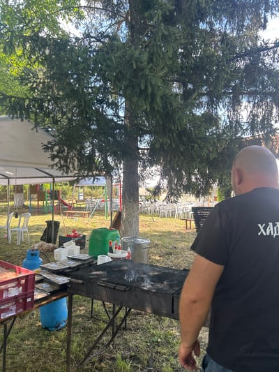 A person wearing a black t-shirt with white text on the back stands near a barbecue grill outdoors. The person is bald and is looking away from the camera. The setting appears to be a backyard or a park, with a large evergreen tree dominating the left side of the frame. In the background, there are several white plastic chairs arranged, and a playground structure is visible. Various items are placed on and around the barbecue grill, including a green plastic container, plastic cups, and what appear to be packages of food wrapped in plastic. A blue propane tank is situated beneath the grill. The ground is covered with grass and dry leaves. The overall scene suggests a casual outdoor gathering or barbecue. The time of day seems to be daytime, possibly late afternoon, given the lighting. The weather appears to be pleasant. The white text on the t-shirt is partially visible and appears to be in a Cyrillic script.
