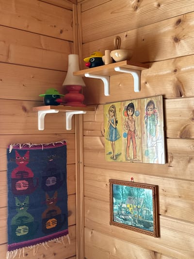 The image shows a corner of a room with wooden plank walls. Two white L-shaped shelves are mounted on the wall. On the upper shelf, there is a ceramic bowl with a handle, a smaller ceramic cup, and a black and yellow toy with red eyes and a red nose. On the lower shelf, there is a black spherical toy with a green hat and red nose, and a red and white ceramic lamp base. To the left of the lower shelf, a woven tapestry featuring stylized cats in various colors hangs on the wall. On the right side of the image, two framed pictures are displayed. The upper picture is a triptych of three young girls in a vintage art style. The lower picture is a framed landscape painting of a yellow house with a water wheel. There is no indication of the city of Borno, Italy, in this image.