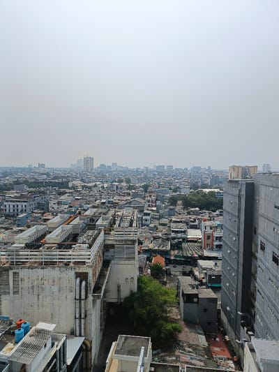 An elevated, wide-angle view depicts a dense urban landscape within the Special Capital Region of Jakarta, Indonesia, under a hazy, pale grey sky. In the immediate foreground, to the left, a large, weathered multi-story building features a flat rooftop crowded with numerous industrial HVAC units and cooling towers, some enclosed in light-colored structures, alongside exposed ductwork and pipes. Vertical grey pipes descend the building's exterior wall, which exhibits discoloration and streaks. Below this, on a lower rooftop, two blue cylindrical water tanks and one orange tank are visible. To the right, a tall, modern, grey building with a grid pattern of dark, rectangular windows dominates the frame. Between these two foreground structures, a dense sprawl of low to medium-rise residential and commercial buildings extends into the mid-ground, characterized by varied rooflines, tight packing, and narrow passages. Sparse green trees are visible in pockets of the mid-ground. The background reveals a distant skyline of numerous high-rise buildings and skyscrapers, partially obscured by the pervasive atmospheric haze. No individuals or discernible activities are present.