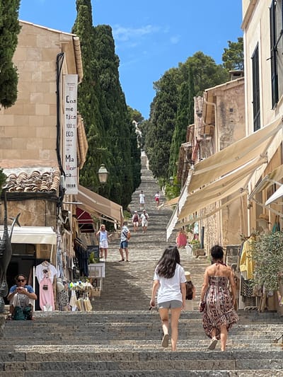 The image depicts a steep, stone staircase in the town of Pollença, Spain, leading up a hillside lined with buildings and cypress trees. The architecture is characteristic of Mediterranean towns, with stone buildings, tiled roofs, and awnings extending over shops. Several people are visible ascending and descending the stairs. On the left side of the stairs, a man sits on the steps, looking at his phone. Shops with clothing and bags are displayed along the lower part of the staircase. Towards the top of the image, the staircase disappears into the distance, framed by dense green trees. The sky is clear and blue, indicating a sunny day. The overall scene suggests a popular tourist destination with a vibrant atmosphere.
