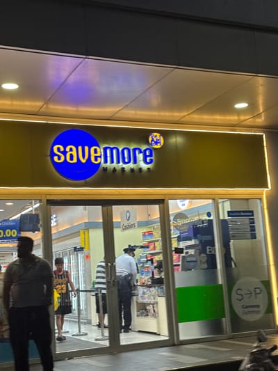 The image shows the exterior of a "Savemore Market" in Manila, Philippines at night. The market has a large, illuminated sign with the words "savemore MARKET" in yellow letters against a golden background. A blue circular logo with yellow lettering is also part of the sign.

Through the storefront windows, several people can be seen inside the market. One person, wearing a white shirt and hat, appears to be standing near the cashier area. Another individual, in a striped shirt and shorts, is visible further inside. In the foreground, the silhouettes of two people are present, suggesting they are customers or passersby. The store interior displays shelves stocked with various products.

Above the entrance, a circular sign with "S→P Currency" is visible, indicating a currency exchange service. The lighting from the market and the street illuminates the scene, reflecting off the building's facade. The overall impression is a bustling retail environment at night. Visible text includes "savemore MARKET", "S→P Currency", and signs within the store such as "Cashiers" and "Premium milk".