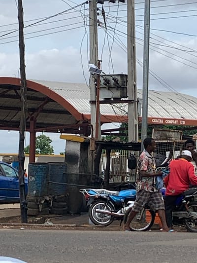 The image depicts an outdoor street scene in Shendam, Nigeria, under an overcast sky with scattered clouds. In the foreground, a dark asphalt road surface is visible.
Two men are present in the mid-ground, standing near motorcycles. The man on the left wears a multi-colored, short-sleeved button-up shirt with a floral or abstract pattern, plaid shorts, and light blue flip-flops. He holds a bundle of light-colored fabric or clothing in his left arm. He is positioned next to a blue motorcycle that appears to be parked, with its front wheel visible. The man on the right wears a red long-sleeved shirt, dark pants, and a white baseball cap. He is partially obscured by the first man and a dark-colored motorcycle, seemingly leaning against or seated on it.
Behind the individuals and motorcycles, to the left, a blue car is partially visible, parked or stopped. Adjacent to it, several dark blue industrial barrels are stacked, with old tires horizontally arranged below them, forming a low barrier or base.
Dominating the mid-ground and background are multiple utility poles and extensive overhead wiring. A thick, dark, possibly wooden pole stands on the far left. Towards the center, a concrete or metal utility pole features a small solar panel and a light fixture attached to its side, along with a transformer or electrical equipment. Numerous black electrical or communication wires crisscross horizontally and vertically between the poles.
In the background, two buildings with distinctive orange-brown arched roof structures and corrugated metal roofing are visible. The building on the right has "ORACHA COMPANY LTD" written in dark letters on its facade. Green foliage and trees are visible between and behind these structures, extending to the horizon.