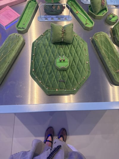 This is a high-angle shot of a jewelry display, featuring multiple green velvet cases showcasing gold jewelry items. The cases are arranged on a metallic surface, with various shapes like rectangles, octagons, and smaller pedestals. Some jewelry pieces are necklaces with pearls and crystals. In the bottom left, there is a pink package and a blue label with the words "SWAROVSKI Ariam Grande CAPSULE COLLECTION". To the right of the scene, a person is visible wearing patterned shoes and a light-colored outfit with silver embellishments. The location is Spreitenbach, Switzerland, based on the provided OCR.
