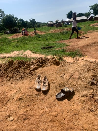 The image depicts a sun-drenched outdoor scene in Buga da Gindi, Nigeria. In the foreground, a pair of worn, light-colored athletic shoes and a black slide sandal are resting on a mound of reddish-brown soil. The ground is a mixture of loose dirt and some dry vegetation. In the middle ground, several people are engaged in activity, some appearing to be working with sand or soil. One woman is visible in the background, carrying a bucket on her head and a blue container in her hand, walking away from the viewer. The environment consists of sparse vegetation, with trees and bushes scattered across the landscape, and a collection of simple structures with corrugated metal roofs in the distance, suggesting a rural or semi-rural settlement. The sky is clear and blue, indicating fair weather.
