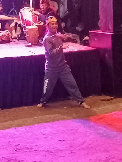 The image captures a vibrant cultural performance, likely an evening event given the artificial lighting, set in Pangalengan, Indonesia. The scene is dominated by a male performer in the foreground, illuminated by purple and pink spotlights.
This man is actively performing, possibly dancing or executing expressive movements, barefoot on a brown, earthy ground. He wears a grey long-sleeved shirt, loose grey pants with an indistinct white graphic or text on the left leg, and a distinctive gold-colored traditional headpiece. He smiles broadly, his hands open and arms bent, suggesting graceful or dynamic motion.
Behind him, on a stage draped in dark fabric, another man is seated and engrossed in playing two kendang, which are traditional Indonesian drums. He also wears a dark top and a similar headpiece. Other instruments or equipment are partially visible on the stage. To the right, dark, stacked rectangular boxes, likely speakers or stage elements, rise beside the stage. A bright pink and red light illuminates a section of the ground in front of the stage. The background is dark and blurred, hinting at an audience or other elements of the venue. The overall impression is one of an engaging and traditional performing arts event.