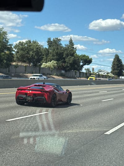 A bright red Ferrari sports car is captured in motion on a sunny day, traveling on a multi-lane highway. The car is the primary subject, positioned in the foreground and viewed from its rear, showcasing its sleek design and prominent exhaust system. Other vehicles are visible on the highway, including a white pickup truck in the middle ground and a yellow sports car further in the distance. The highway is bordered by a concrete barrier and lush green trees, suggesting a suburban or semi-rural environment. In the background, a residential house with a light-colored exterior is visible among the trees. The sky is a clear blue with scattered white clouds, indicative of fair weather. The scene appears to be a typical highway commute in Roseville, USA, during daylight hours.