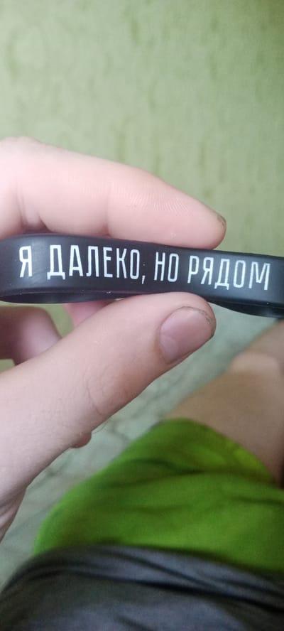 The image shows a close-up of a person's hand holding a black silicone wristband with white Cyrillic text that reads "Я ДАЛЕКО, НО РЯДОМ" (Ya daleko, no ryadom), which translates to "I am far, but near." The background is a blurry green textured surface, possibly a wall or fabric. The wristband is being held by the thumb and forefinger, obscuring some of the text. The hand appears to be that of an adult, with visible fingerprints and a fingernail. The image quality is moderate, with some graininess. No other individuals or objects are clearly visible. The image does not provide any information to determine its location in Kudymkar, Russia.