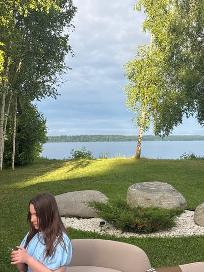 The image depicts a serene natural landscape, likely near the city of Osipovo, Russia, on a cloudy day. In the foreground, a young girl with long brown hair, wearing a light blue dress, is seated on a beige sofa, engrossed in her mobile phone. The immediate surroundings are a lush green lawn, with two large, grey, weathered rocks and a small evergreen bush placed on a bed of white pebbles.

Beyond the lawn, a vast body of calm water stretches towards a distant, tree-covered shoreline. The water reflects the soft, diffused light of the overcast sky. Several birch trees with white bark are scattered across the landscape, their green leaves casting dappled shadows on the grass, indicating the direction of the sun. The overall atmosphere is one of peacefulness and quietude, with the natural elements dominating the scene.