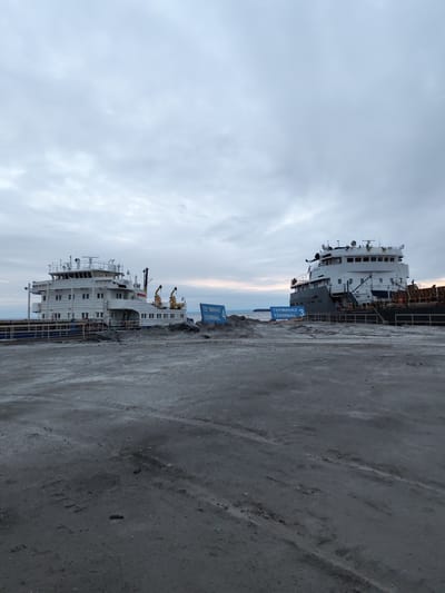 The image depicts a port area in Shcheleiki, Russia, under an overcast sky. Two large vessels are docked at Terminal 3 and Terminal 4, respectively. The ground appears to be covered in a gray, sandy material, possibly cement. The vessels are predominantly white and gray, with visible cranes on the vessel docked at Terminal 4. The signs indicating the terminals are blue and white. The scene is still, lacking apparent human activity. The image suggests an industrial or commercial port environment.