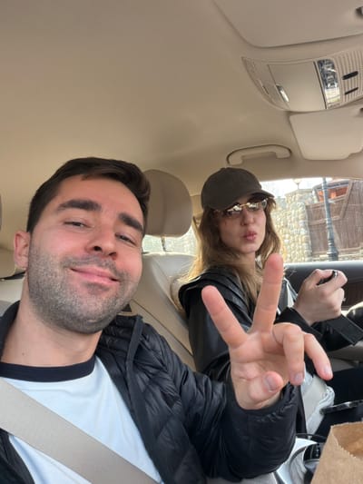 The image shows two people in a car. The man in the foreground is wearing a white t-shirt with black trim, a black jacket, and a seatbelt. He is looking at the camera. Behind him, a woman is wearing a black jacket, a black cap, and sunglasses. She is holding up her fingers in a V-sign. The interior of the car is light-colored. The location is identified as Selyatino, Russia, based on the presence of a building that appears to be built out of stone that can be seen through the window.