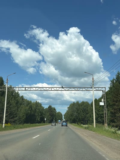 A vehicle is driving on a paved road with a clear blue sky and white clouds above. The road is lined with trees on both sides, and there is a metal structure spanning the road with traffic lights or signs attached. There are other vehicles visible on the road ahead. The environment appears to be a rural or semi-rural area, possibly on the outskirts of Kamennoye, Russia, given the presence of dense forestation and the style of infrastructure. The lighting suggests it is daytime.