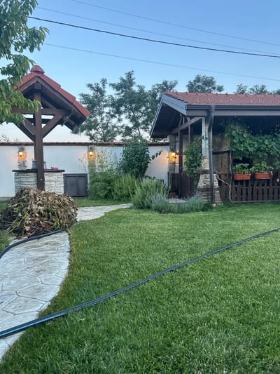 The image depicts a backyard scene in the village of Voyvodino, Bulgaria. The focal points are a covered outdoor kitchen or grill area with a red tile roof and stone accents, and a smaller wooden structure with a similar red tile roof, possibly a well cover or gazebo. A stone pathway winds through a well-maintained green lawn. To the left of the pathway, a large pile of dried leaves and branches is visible. In the background, a white wall with two wall-mounted lights is present, along with lush green trees and shrubs. The sky is clear and blue, suggesting it is either early morning or late evening. There are no people or animals visible in the image. A black hose lies on the grass, partially crossing the stone path.