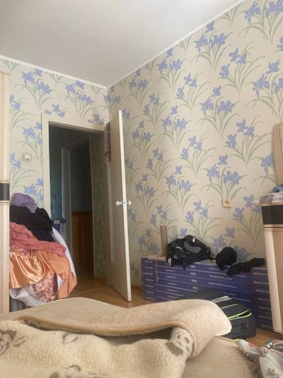 The image depicts an interior scene within what appears to be a room in Kovrov, Russia. The walls are adorned with a pale cream wallpaper featuring a pattern of blue irises and green leaves. To the left, a portion of a wooden wardrobe is visible. In the foreground, a pile of folded clothes, including items in orange, pink, and dark purple, is situated near a doorway that leads into another room. The doorway itself is open, revealing a glimpse of a hallway with green floral wallpaper and wooden paneling.

On the right side of the frame, a closed white door with a doorknob is partially open, suggesting it leads to another part of the residence. Near this door, on the wall, is a light-colored electrical outlet. Further to the right, a wooden dresser or cabinet stands against the wall.

On the floor, a stack of blue boxes, possibly containing flooring material labeled "QUICK-STEP LAMINATE," is visible. On top of these boxes, a black backpack and a black hat are placed. In the immediate foreground, a beige blanket with a textured pattern is draped over what appears to be a rolled-up rug or another blanket. The overall impression is of a lived-in domestic space.