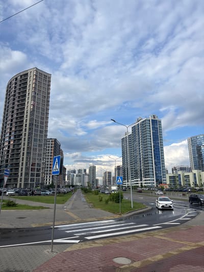 The image captures a street scene in the Senitsky Sel'skiy Sovet, Belarus, dominated by modern, high-rise apartment buildings. On the left, a distinctive curved, grey building with numerous windows stands prominently. To the right, two taller, slender buildings with blue and white facades are visible, with more buildings stretching into the distance. The sky is overcast with a mix of white and grey clouds. A wet asphalt road curves through the scene, with a white car turning. A pedestrian crossing with white stripes is in the foreground, with red and grey paving stones. Traffic signs, including a blue sign with a white pedestrian symbol, are present. Cars are parked along the roadside, and a few pedestrians are seen walking on a sidewalk. The overall impression is an urban environment with contemporary architecture under a cloudy sky.