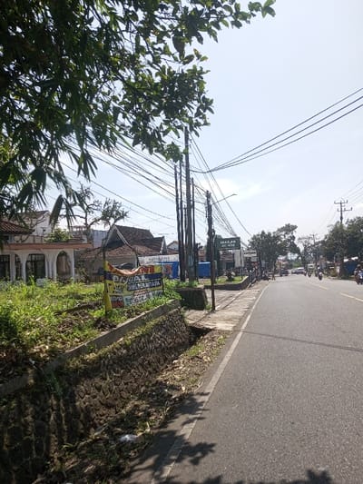 The scene depicts a sunny, daytime view along a semi-urban road, identified as Beteng_Pangkhar_Somthrang, Bhutan. A two-lane asphalt road occupies the center and right foreground, extending towards the upper right background. Shadows from overhead foliage are cast across the road's foreground.
On the left side of the road, a concrete sidewalk is present, bordered by a stone-faced drainage ditch that contains dried leaves and some debris. Above the ditch and sidewalk is an elevated patch of unkempt, overgrown grass. A prominent yellow and blue banner sign is displayed in this grassy area, facing the road. The sign features text in Indonesian, including "DIJUAL" (For Sale), "TANPA PERANTARA" (No Middleman), and "2.5M". Behind this sign, several buildings are visible; the closest is light-colored with arched openings, while others further back have pitched, tiled roofs. A dense array of utility poles, some wooden and others concrete, line the left side of the road, supporting a complex network of overhead electrical and communication wires. Lush green foliage, likely from trees or bamboo, partially obscures the upper left quadrant of the image.
On the right side of the road, in the mid-ground, additional utility poles are visible. Several commercial structures are present, one with a sign partially legible as "MASJID AL-IMAN" and another suggesting "MATAHARI." Multiple motorcycles, some with riders, are observed moving away from the camera or parked along the road's edge. A light-colored vehicle is also visible further down the road. The sky above is largely clear with a light blue hue.