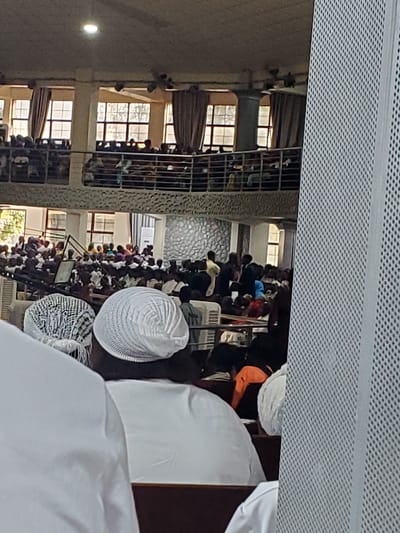 This media file shows a gathering of Muslim men inside what appears to be a mosque in Kason, Nigeria. The image is taken from a high angle, looking down on the congregants. The men are dressed in traditional attire, primarily white robes and caps, and are engaged in prayer. Some are standing, while others are in a kneeling or prostrating position, indicating the prayer is in progress.
The setting features architectural elements typical of a mosque, including windows with decorative grilles and textured stone walls. The lighting suggests it could be daytime, possibly late afternoon or early evening, as there is natural light filtering in from the windows. The overall atmosphere appears solemn and focused, consistent with a religious service. The presence of multiple rows of people praying together signifies a communal prayer session.
Notable details include the variety of kufi caps worn by the men, ranging from simple white ones to more intricately patterned designs. There is a sense of community and shared devotion among the individuals. The image is a snapshot of a religious event, capturing a moment of collective worship. No visible text is present in the image.
