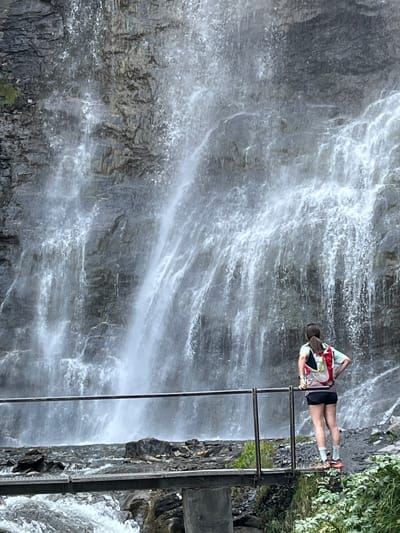 A person stands on a metal bridge, facing a tall waterfall that cascades down a rocky cliff. The person, viewed from behind, wears a light green shirt, black shorts, white socks, and athletic shoes. They have a backpack with red, white, and yellow accents. A metal railing runs along the bridge and the person's right hand rests on it. The waterfall is the dominant feature, with white water flowing over grey rock. The ground around the base of the waterfall is rocky, with some green vegetation visible to the right. The water below the bridge is turbulent and white, indicating a fast current. The scene appears to be an outdoor natural setting. The image is likely taken in the vicinity of Benasque, Spain, given the presence of a waterfall and mountainous terrain common to the region.