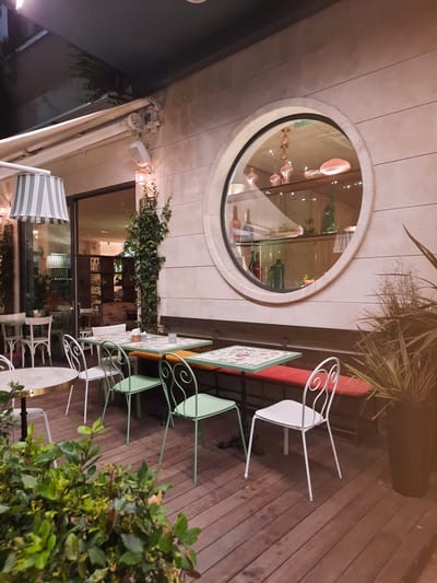 The image shows an outdoor seating area of a restaurant or cafe. The floor is made of wooden planks, and there are several tables with chairs. Some tables are covered with cloths decorated with floral patterns, while others appear to have plain surfaces. The chairs are of various designs, including white metal chairs with decorative backs and light green metal chairs.

In the background, there is a wall made of beige stone blocks. A large, circular window is set into the wall, and inside the window, shelves are visible displaying various glass bottles and decorative objects. A vine with green leaves is growing up the wall next to the window.

To the left of the circular window, there is a glass door leading into the establishment, revealing more seating and shelves inside. A light fixture with a striped lampshade hangs from an awning above. In the foreground, there is a large potted plant with spiky leaves and a lush green bush. The lighting suggests it is evening or a dimly lit interior space. The overall scene depicts a cozy and inviting outdoor dining environment. The image is likely from Pesaro, Italy, based on the prompt's request.