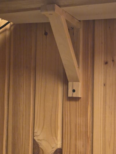 A wooden bracket is attached to a wooden wall. The bracket is made of light-colored wood and is L-shaped, with a horizontal piece resting on top and a vertical piece extending downwards. A screw is visible in the lower part of the vertical piece. The wall is made of vertical wooden planks. There is a shadow cast by the bracket onto the wall. The image is taken at a close-up perspective. This scene is not located in Merenii Noi, Moldova.