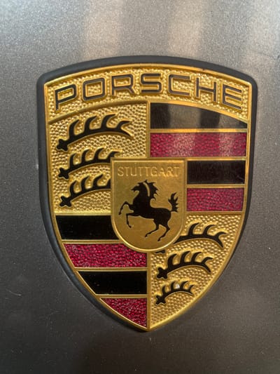 The image shows a close-up of a Porsche crest emblem, likely affixed to a vehicle. The emblem is shield-shaped with a gold border and black outer trim. The upper portion of the crest features the word "PORSCHE" in raised gold lettering against a black background. The central part of the crest contains the Stuttgart coat of arms, which includes a black prancing horse on a gold background, and the word "STUTTGART" in gold lettering above the horse. The crest is divided into quadrants, with alternating sections of gold textured with a honeycomb pattern and black and red stripes. The red stripes have a glittery texture. The emblem is mounted on a dark gray, slightly textured surface, possibly the body of a car. There are no people visible in the image. The image is taken from a straight-on perspective, with no specific location in Gerstheim, France, identifiable from the emblem itself.