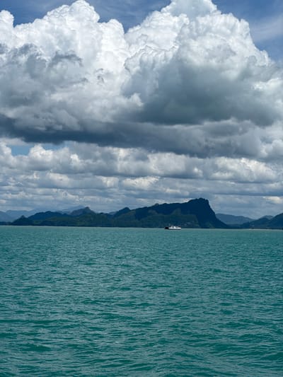 The image shows a vast expanse of turquoise ocean under a dramatic sky filled with large, fluffy cumulus clouds interspersed with darker, more ominous storm clouds. In the distance, a rugged, tree-covered island chain forms a dark silhouette against the sky. A small white ferry boat with red accents is visible in the middle distance, navigating the calm waters. The foreground is dominated by the textured surface of the ocean, with small waves creating ripples and shadows. The overall scene evokes a sense of natural beauty and scale, with the contrasting elements of the stormy sky and serene water. The location is identified as Thong Nian, Thailand.