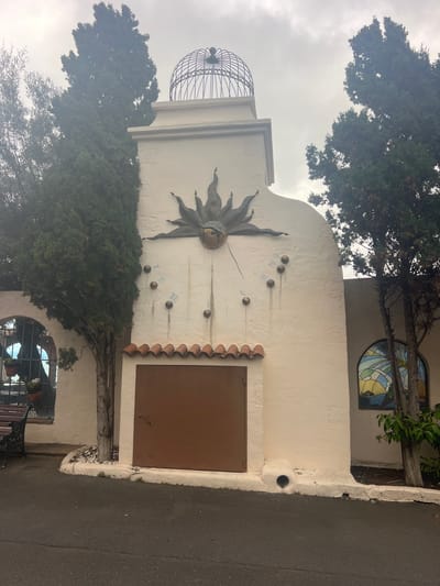 The image shows an outdoor scene in the city of La Vera, Spain. The focal point is a cream-colored stucco building with a large, ornate sundial featuring a sunburst motif. The sundial has Roman numerals and several small, spherical hour markers. Above the sundial is a decorative metal dome. To the left of the sundial, a tall, dark green evergreen tree stands close to the building. A wrought-iron bench with a wooden seat is visible in the lower left corner, with an arched window behind it containing potted plants. To the right of the sundial, another evergreen tree is present, and a stained-glass window with a colorful, abstract design is set into the wall. A brown metal door is positioned below the sundial. The ground in front of the building is paved with asphalt. The sky is overcast, suggesting a cloudy day.