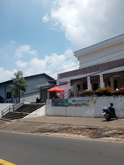 A street scene shows a building identified by a sign as "Masjid Sarah," a mosque, in daylight. The sky is blue with scattered clouds. A motorbike is parked in front of the mosque. A red canopy is visible near a staircase leading to another building next to the mosque, and there appears to be people seated beneath the canopy. Banners with the words "IDUL FITRI" are attached to the wall. The location is not in Nawng Hkar, Myanmar.