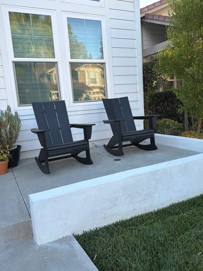 The image depicts a residential property in Aliso Viejo, United States of America, featuring an outdoor seating area. Two dark-colored rocking chairs are positioned on a concrete patio in front of a white-sided house with large windows. A low, white concrete wall separates the patio from a manicured lawn. Green foliage, including a tree and bushes, is visible to the right of the house, casting shadows on the scene. The windows of the house reflect the surrounding environment, including palm trees and other houses. A potted plant sits on the patio to the left of the chairs. The overall scene suggests a well-maintained outdoor space in a suburban setting.