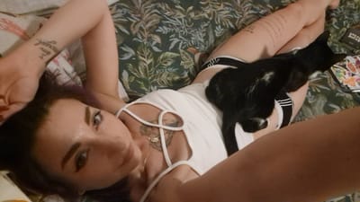 A woman is lying on a bed with a black and white cat on her. The woman has dark hair and is wearing a white top and black and white striped underwear. She has tattoos on her arm and chest. The cat is lying on her stomach and chest. The bed is covered with a floral patterned duvet. There is a book with the words "BREAK RULES" on it on the bed to the right of the woman. The image appears to be taken from a high angle, looking down at the woman and cat. The scene is indoors. There is no information that indicates the location is in Reus, Spain.
