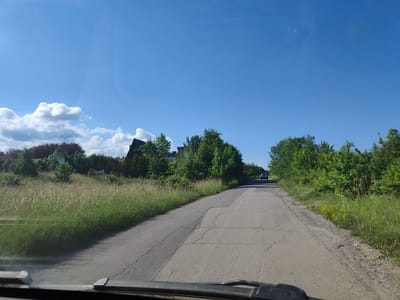 The image shows a view from inside a vehicle, looking forward along a paved road. The road is cracked and appears aged. On the left side of the road is a grassy field with tall grass, and beyond that, there are trees and the roofs of buildings, suggesting a suburban or rural setting. On the right side of the road, there is a dense line of green trees. The sky is clear and blue with a few scattered white clouds. In the distance, a silver car is visible driving away from the camera on the same road. The scene is likely in the city of Rudartsi, Bulgaria, given the context provided, and appears to be a sunny day.