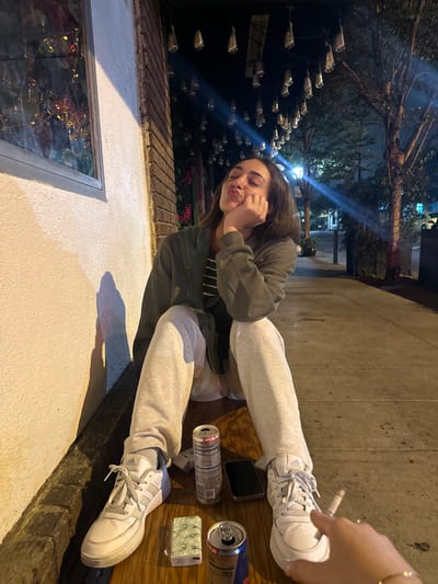 The scene is located in Glendale, United States, at night. A young adult female subject is seated on a light-colored wooden surface, leaning against a building wall. She has brown hair and is dressed in a dark grey zip-up hoodie, a striped shirt, light grey sweatpants, and white Adidas brand sneakers. Her left elbow rests on her left knee, with her left hand supporting her chin. Her eyes are partially closed, and her lips are pursed. A shadow is cast on the wall to her left.
To the subject's left, the building wall features a light-colored lower section transitioning to brick, with a window reflecting multi-colored lights. Overhead, numerous bare incandescent light bulbs are strung, forming a canopy. In the background, trees, a street, and urban structures are dimly visible under artificial streetlights.
On the wooden surface in front of the female subject, the following objects are present: an open silver and red Red Bull energy drink can, another open silver, pink, and purple beverage can with a nutrition facts label, a black smartphone with a dark screen, and a small, light-colored rectangular object with a repeating green pattern. In the lower right foreground, a right hand, adorned with a bracelet, is visible holding a lit cigarette, indicating the presence of a second individual.