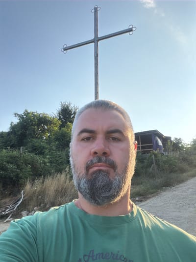 A man, with a greying beard and hair, is taking a selfie outdoors. He is wearing a green t-shirt. In the background, a large metal cross stands against a clear blue sky. To the right of the cross, there is a small, rustic structure covered with a blue tarp. The environment appears to be a rural or semi-rural area, with trees and dry grass visible. The lighting suggests it is either early morning or late afternoon. The scene is located in Gniazdovo, Bulgaria.