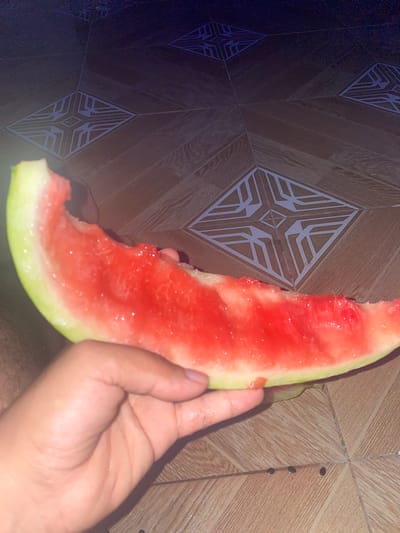 A hand holds a partially eaten slice of watermelon over a tiled floor. The watermelon has a green rind and bright red flesh, with some seeds visible. The floor tiles are a light brown with a repeating geometric pattern in a lighter color. The background is dark and out of focus, suggesting low lighting conditions. The image appears to have been taken indoors, possibly in a residential setting. There are no identifying landmarks or indications of a specific city location.