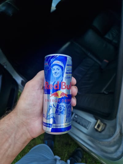 A close-up, eye-level shot captures a person's hand holding a Red Bull energy drink can, presented at a slight upward angle in front of the interior of a vehicle. The can is predominantly blue and silver with graphics of female athletes and the Red Bull logo. The vehicle's interior, visible in the background, appears to be dark, possibly leather seats, and a silver door frame. The scene suggests an outdoor setting due to the presence of greenery and grass at the bottom of the frame. The image appears to be taken on a sunny day.