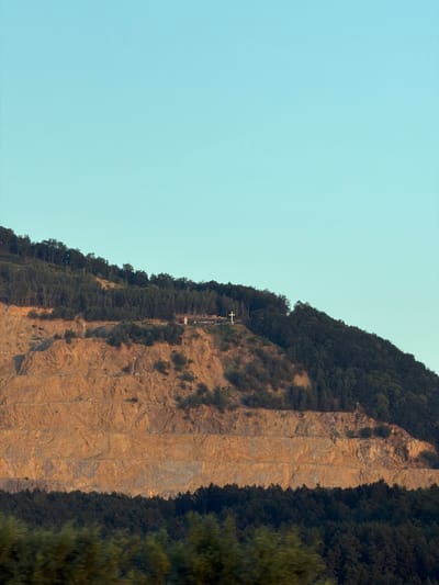 This media file depicts a large, steep, rocky cliff face covered in dense green trees along its upper edges. The cliff itself is a striated, light brown color, suggesting sedimentary rock. A thin, white cross is visible on the cliff face, situated on a ledge partway up, accompanied by a series of small, silhouetted structures or lights, possibly representing a pilgrimage path or religious monument. The sky is a clear, pale blue, indicating daylight, likely late afternoon or early morning due to the warm lighting on the rock face. The overall scene is one of natural grandeur with a touch of human religious devotion. The image is oriented vertically, with the sky occupying the left two-thirds and the cliff face and trees filling the right third. There is no visible text in the image. This scene is located in Ladce, Slovakia.