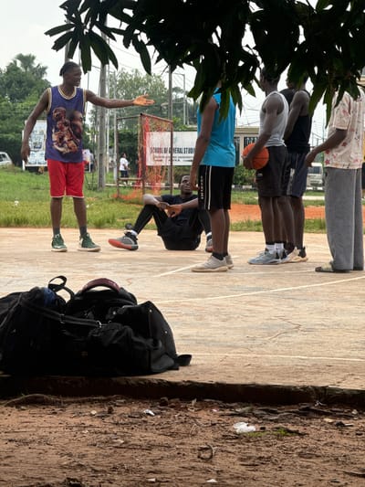 In Amansee, Nigeria, an outdoor basketball court is visible, with several young men gathered. In the foreground, two large black duffel bags are placed on the ground. A man wearing a purple sleeveless jersey with images of basketball players and red shorts stands with his left arm extended, palm open, in a speaking gesture. Next to him, another man is seated on the court wearing black athletic attire.
Behind the seated man, a group of four men are standing in a line. The first man from the left is wearing a blue sleeveless shirt and black shorts. The second man holds an orange basketball and wears a grey sleeveless shirt and black shorts. The third man is in a black sleeveless shirt and dark shorts. The fourth man wears a patterned short-sleeved shirt and grey sweatpants.
In the background, a sign reads "SCHOOL OF GENERAL STUDIES" and mentions "NNAMDI AZIKIWE UNIVERSITY, AWKA". Trees and other greenery surround the court, and the sky is overcast. A white vehicle is visible in the far background. The court surface appears to be concrete with visible lines. Debris, including plastic wrappers and plant matter, is scattered on the dirt area bordering the court.