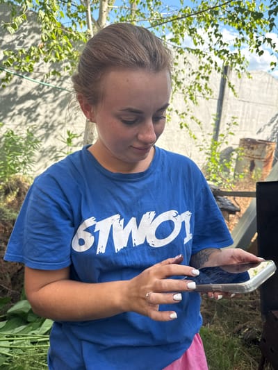 The image shows a young person with blonde hair and fair skin, looking down at a smartphone. They are wearing a blue t-shirt with white text that appears to read "SMOOT." The person has freckles on their face and white nail polish on their fingers. A silver ring is visible on their left hand.
The setting appears to be outdoors, with greenery and a concrete wall in the background. Sunlight filters through the leaves of a tree, suggesting it is daytime. The sky is visible and appears to be blue with some clouds. The overall mood is casual and relaxed.
Notable details include the person's engagement with their phone, the visible text on their shirt, and the outdoor environment. The image is oriented vertically.