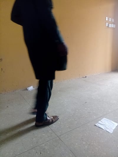 This indoor image, taken in Gauta, Nigeria, shows a partial view of a person in motion. The individual is seen from the mid-torso downwards, with their upper body heavily blurred, indicating movement. They are dressed in dark trousers or a traditional long garment and are wearing distinctive brown monk strap shoes with multiple gold-colored buckles on the side. A long, dark shadow stretches from the person's feet across the tiled floor.

The room features light yellow or beige walls that appear somewhat worn, with minor stains and scuff marks. On the wall to the right, a cluster of white rectangular light switches or power outlets is visible. The floor consists of light grey or off-white tiles, possibly terrazzo, with visible grout lines. Two crumpled pieces of white paper or tissue are scattered on the floor: one near the base of the wall close to the person, and another further out in the foreground. The lighting suggests an artificial source, casting the prominent shadow. No discernible text is visible anywhere in the image.