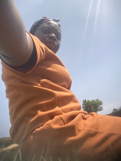 The image, taken in Kukum, Nigeria, presents a low-angle perspective of a person with dark skin looking directly into the camera. The individual is positioned against a bright, clear blue sky, suggesting an outdoor setting under strong sunlight.
The subject, likely female, wears a loose-fitting, long-sleeved top and what appears to be matching bottoms in a warm orange-brown color. The fabric exhibits a soft, slightly textured quality, possibly fleece or thick cotton, with visible seams and folds indicating a relaxed fit. A dark, patterned head covering or bandana is worn around the head. The subject's face is angled slightly downwards towards the camera, displaying a calm, neutral, or subtly pensive expression. Highlights on the skin are noticeable, consistent with direct sunlight.
In the background, a portion of a lush green tree with dense foliage is visible in the lower part of the frame (when the image is viewed upright). The vast expanse of clear blue sky fills most of the background, with some soft light flares indicative of bright daylight. The scene captures a casual, sunlit moment outdoors. No text is visible in the image.