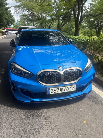 A blue BMW is parked on an asphalt road in Gyeongju-si, South Korea. The car is facing forward, and its front grille, headlights, and license plate are visible. The license plate reads "267부 6714". Behind the BMW, other vehicles are parked along the road, including a white car and a bus. The scene is surrounded by lush green trees and bushes, suggesting a natural or park-like setting. The sky is partly cloudy.