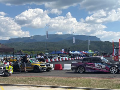 In Dospey, Bulgaria, on a sunny day with scattered clouds, a motorsport event is underway. The image captures a section of a race track with several drift cars parked or in motion. In the foreground, two prominent cars are visible: a grey and yellow BMW adorned with racing decals and a black BMW with a vibrant pink and purple graphic wrap that reads "K&P AUTOMATIC TRANSMISSION SERVICE." Several individuals are present around the cars, some appearing to be participants or crew members. One man in a yellow safety vest is kneeling by a backpack, while another man in a black t-shirt is leaning into the grey and yellow BMW. In the background, a row of tents and banners suggests a larger event setup, with spectators gathered behind safety barriers. Beyond the track, a dense forest covers the foothills of imposing mountains, with ski slopes visible on one of the peaks. Streetlights and flags are also scattered throughout the scene, indicating the race circuit's infrastructure.