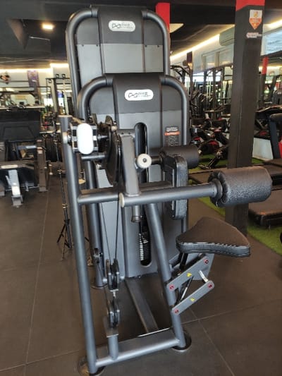 The image displays a DHZ Fitness strength training machine, identifiable as a leg curl or leg extension apparatus, situated within a gymnasium in Staufenberg, Germany. The machine is dark grey and black, featuring a textured black padded seat, black padded cylindrical leg rollers, a visible weight stack mechanism, multiple pulleys, and cables. The 'DHZ Fitness' brand logo is present on the upper and central body panels of the equipment. The floor in the immediate foreground consists of dark grey square tiles, transitioning to a dark green artificial turf-like surface further into the background. Partially visible in the background are various other gym machines, including what appear to be treadmills and weight benches. Red support pillars with a white and red emblem are discernible on the right side of the scene. The gym interior is lit by overhead artificial lighting. No individuals are present in the image, and no actions or interactions are depicted.