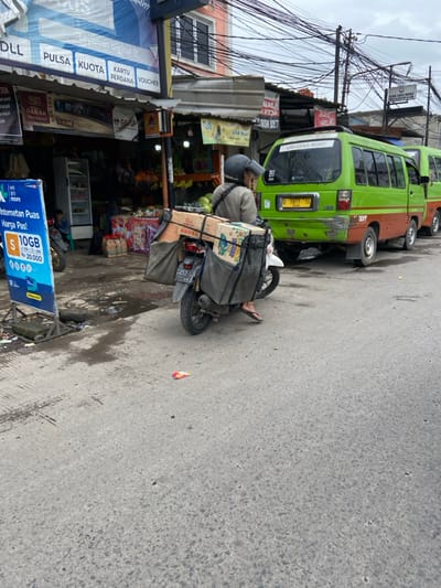 The scene is on a street in Kecamatan Dayeuhkolot, Indonesia, with a man on a motorcycle that is loaded with boxes. He is wearing a helmet and a long-sleeved shirt. The motorcycle is facing the right of the frame, and is parked at the edge of the street. There is a green van parked next to the motorcycle. The van is facing the opposite direction. The van is in the middle of the street, and is parked next to a building. The building has a sign that says "Jual Aksesoris". The van has a license plate that reads "D 1977 YG". There is a sign on the left side of the frame that advertises a mobile phone plan. The street is paved and wet. The sky is overcast. The man is wearing sandals. There are power lines and wires strung overhead. There are at least 2 other people on the sidewalk behind the motorcycle. There is a pile of boxes stacked near the motorcycle. The man is holding a brown bag on his right shoulder. The scene is during the daytime.