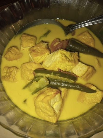 The image presents a close-up, overhead view of a prepared food dish contained within a clear, fluted serving bowl. This scene is located in the city of Doramamidi, India. The bowl holds a yellow, opaque curry or gravy base. Submerged in the liquid are numerous irregular, roughly square-shaped pieces of light yellow-brown fried tofu or paneer. Several dark green chili peppers, some appearing longitudinally split, and scattered reddish-orange fragments, possibly tomatoes, are also visible among the solid ingredients. A silver-colored metal spoon, featuring an ornate handle design, is partially submerged in the upper right quadrant of the bowl. No individuals are present in the frame. The lighting appears to be artificial, highlighting the textures and colors of the dish.