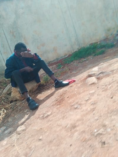 A young man, dressed in a dark blue jacket and black pants, is sitting on a rock. He is looking down, his left hand covering his face. He is wearing black shoes and a red shirt is visible underneath his jacket. There is a large rock behind him and the ground is dirt. He appears to be sitting on the edge of an unpaved road or path. There is short green grass in the background. This scene is located in Gitaru, Kenya, at coordinates -1.218, 36.655.