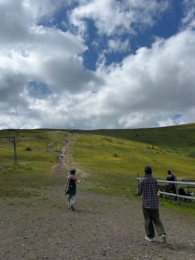 In Chorna Tysa, Ukraine, this image depicts a grassy mountain slope with a dirt path leading upwards. Several people are seen walking along the path. In the foreground, a person in a plaid shirt and dark pants walks away from the camera. To the left, another person with a backpack walks further up the path. In the background, ski lifts are visible on the slope. The sky is partly cloudy with a mix of blue sky and white clouds. A wooden fence with seating is present on the right side of the frame.