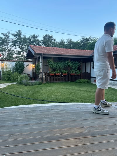 A full shot shows a man in a white polo shirt and white shorts, wearing athletic shoes, standing on a wooden deck and looking to the right. Behind him, a green lawn leads to a garden with a thatched-roof structure, a white wall, and trees. The sky is clear and blue, with visible power lines. The scene appears to be in a backyard in the village of Voyvodino, Bulgaria.