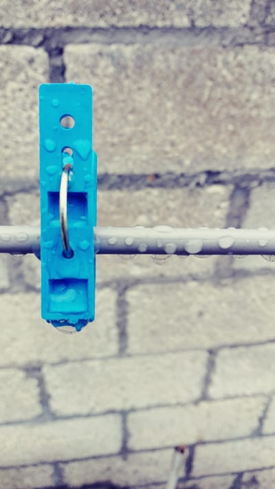 This image, captured in the city of La Joya, Mexico, presents a close-up, vertical perspective of a blue plastic clothespin. The clothespin is prominently featured, centrally positioned, and exhibits multiple small, reflective water droplets adhering to its surface. A larger, spherical water droplet is suspended from the lower jaw of the clothespin. It is clipped onto a horizontal, light-grey cylindrical rod or line, which is also covered in numerous small water droplets across its visible length. The background consists of an out-of-focus, light-colored concrete block wall, displaying blurred darker lines consistent with mortar joints. No people or other distinct objects are present in the scene.